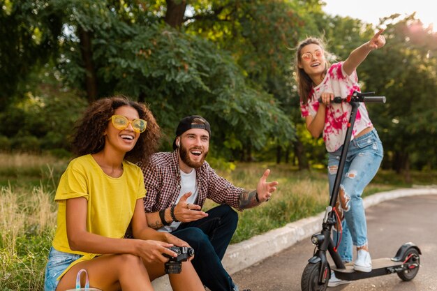 Glückliche junge Gesellschaft von lächelnden Freunden, die im Park auf Gras mit elektrischem Tretroller sitzen, Mann und Frauen, die Spaß zusammen haben