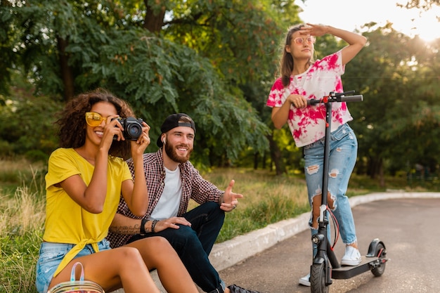 Glückliche junge Gesellschaft von lächelnden Freunden, die im Park auf Gras mit elektrischem Tretroller sitzen, Mann und Frauen, die Spaß zusammen haben