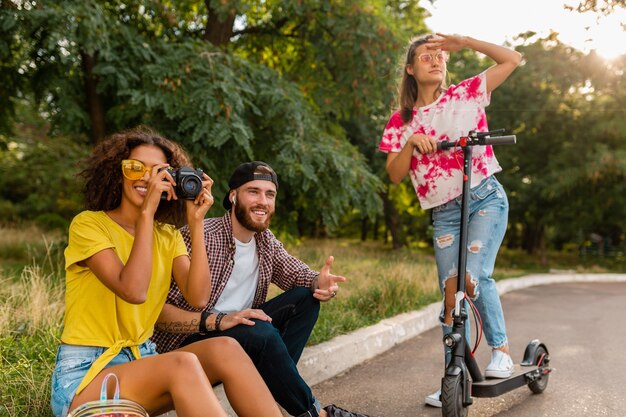 Glückliche junge Gesellschaft von lächelnden Freunden, die im Park auf Gras mit elektrischem Tretroller sitzen, Mann und Frauen, die Spaß zusammen haben