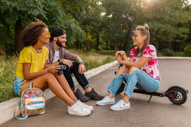 Glückliche junge Gesellschaft von lächelnden Freunden, die im Park auf Gras mit elektrischem Tretroller sitzen, Mann und Frauen, die Spaß zusammen haben