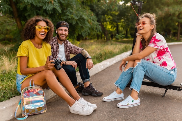 Glückliche junge Gesellschaft von lächelnden Freunden, die im Park auf Gras mit elektrischem Tretroller sitzen, Mann und Frauen, die Spaß zusammen haben