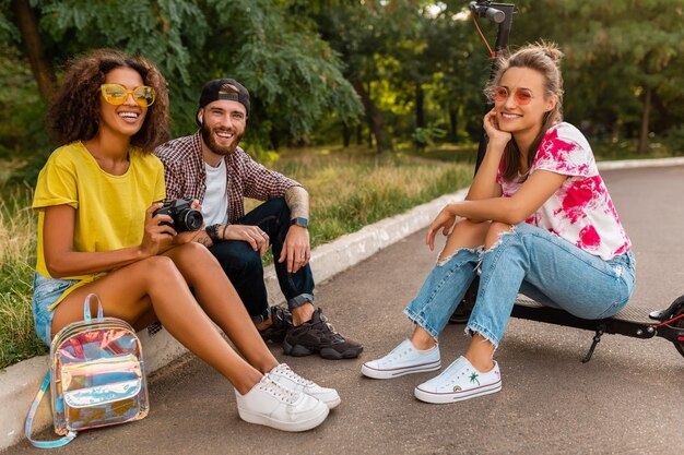 Glückliche junge Gesellschaft von lächelnden Freunden, die im Park auf Gras mit elektrischem Tretroller sitzen, Mann und Frauen, die Spaß zusammen haben