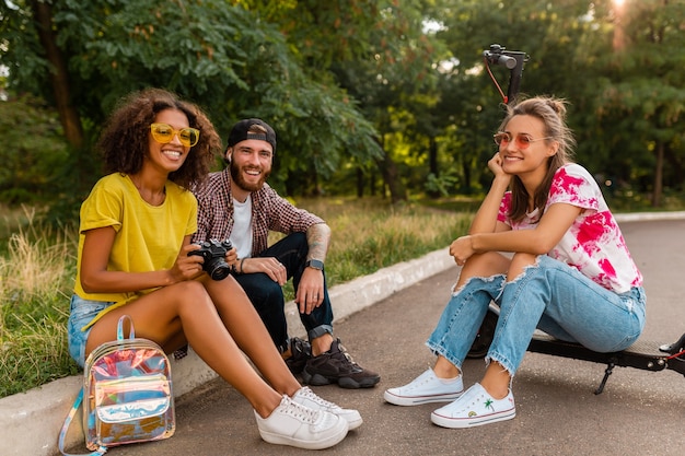 Glückliche junge Gesellschaft von lächelnden Freunden, die im Park auf Gras mit elektrischem Tretroller sitzen, Mann und Frauen, die Spaß zusammen haben