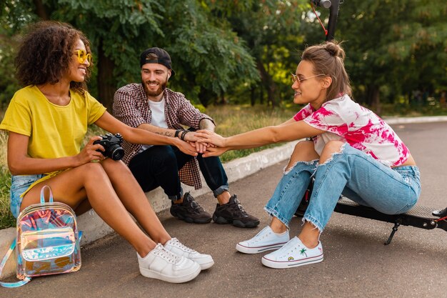 Glückliche junge Gesellschaft von lächelnden Freunden, die im Park auf Gras mit elektrischem Tretroller sitzen, Mann und Frauen, die Spaß zusammen haben