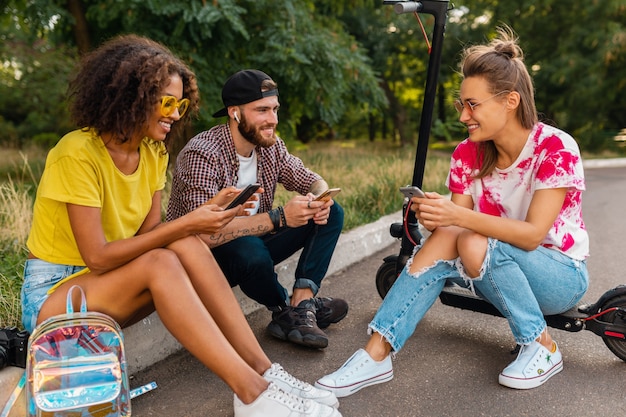 Glückliche junge Gesellschaft von lächelnden Freunden, die im Park auf Gras mit elektrischem Tretroller sitzen, Mann und Frauen, die Spaß zusammen haben