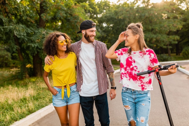 Glückliche junge Gesellschaft von lächelnden Freunden, die im Park mit elektrischem Tretroller, Mann und Frauen gehen, die Spaß zusammen haben