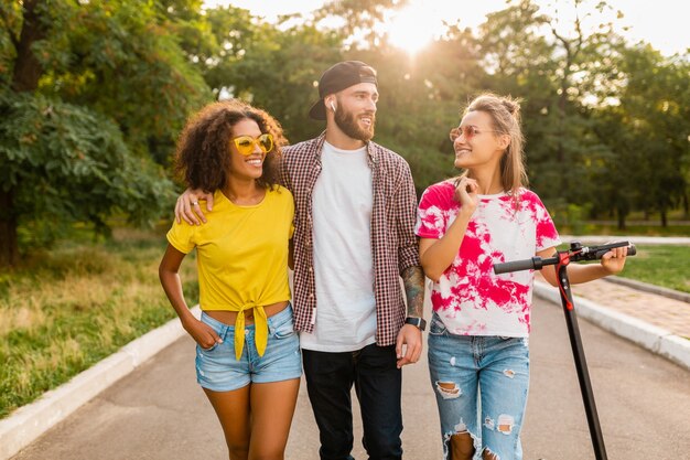 Glückliche junge Gesellschaft von lächelnden Freunden, die im Park mit elektrischem Tretroller, Mann und Frauen gehen, die Spaß zusammen haben