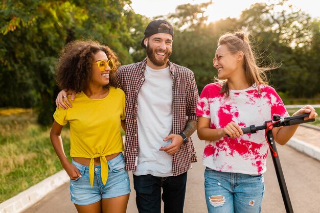 Glückliche junge Gesellschaft von lächelnden Freunden, die im Park mit elektrischem Tretroller, Mann und Frauen gehen, die Spaß zusammen haben