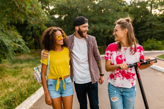 Glückliche junge Gesellschaft von lächelnden Freunden, die im Park mit elektrischem Tretroller, Mann und Frauen gehen, die Spaß zusammen haben