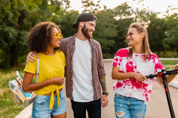 Glückliche junge Gesellschaft von lächelnden Freunden, die im Park mit elektrischem Tretroller, Mann und Frauen gehen, die Spaß zusammen haben