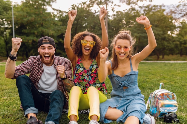 Glückliche junge Gesellschaft von lächelnden Freunden, die Park auf Gras sitzen, Mann und Frauen, die Spaß zusammen haben