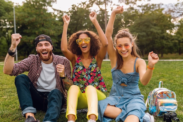 Glückliche junge Gesellschaft von lächelnden Freunden, die Park auf Gras sitzen, Mann und Frauen, die Spaß zusammen haben