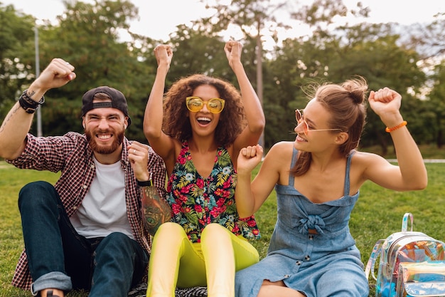 Glückliche junge Gesellschaft von lächelnden Freunden, die Park auf Gras sitzen, Mann und Frauen, die Spaß zusammen haben