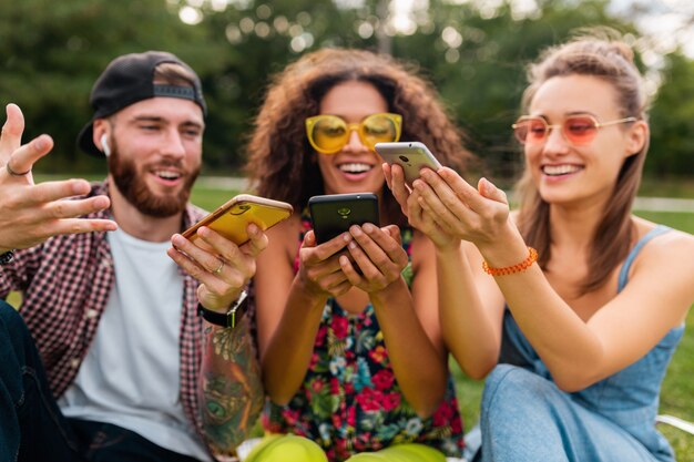 Glückliche junge Gesellschaft von lächelnden Freunden, die Park mit Smartphones, Mann und Frauen sitzen, die Spaß zusammen haben