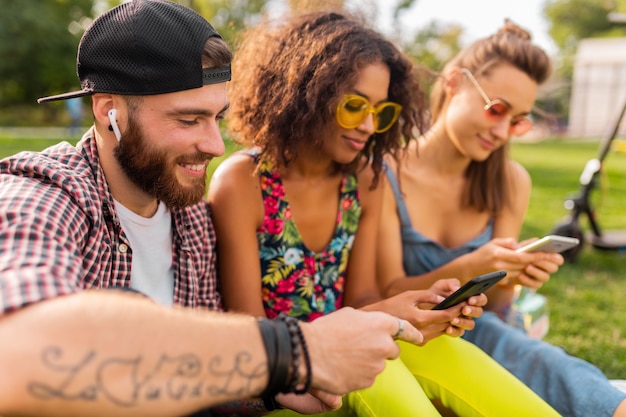 Kostenloses Foto glückliche junge gesellschaft von lächelnden freunden, die park mit smartphones, mann und frauen sitzen, die spaß zusammen haben