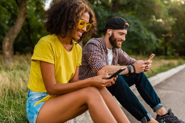 Glückliche junge lächelnde Freunde sitzen Park mit Smartphones, Mann und Frau, die Spaß zusammen haben