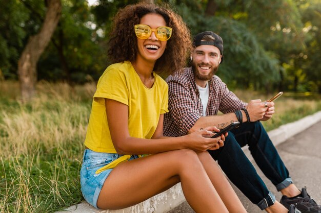 Glückliche junge lächelnde Freunde sitzen Park mit Smartphones, Mann und Frau, die Spaß zusammen haben