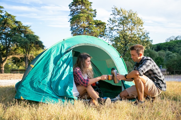 Glückliche junge Leute, die auf Rasen kampieren und im Zelt sitzen. Zwei Reisende und Freunde, die Tee vom Vakuumkolben trinken. Frau, die Stange hält. Backpacking Tourismus, Abenteuer und Sommerurlaub Konzept