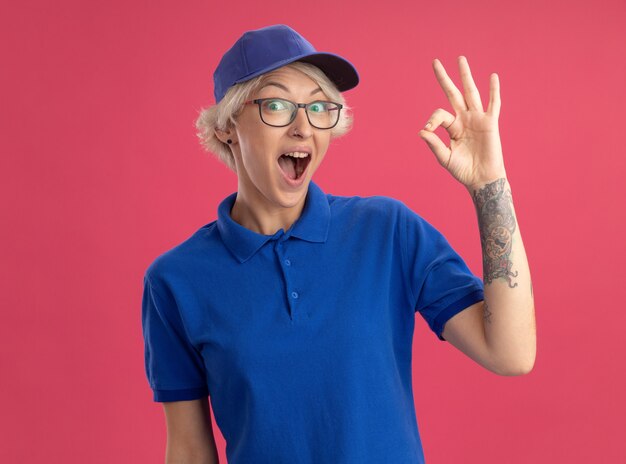 Glückliche junge Lieferfrau in blauer Uniform und Mütze überrascht und erstaunt, ok Zeichen über rosa Wand zeigend