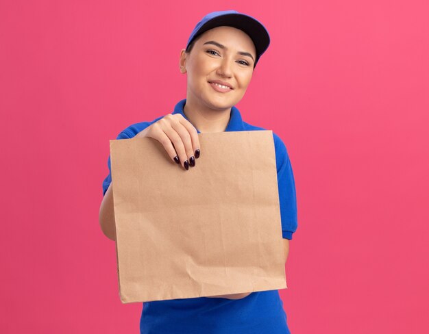 Glückliche junge Lieferfrau in der blauen Uniform und in der Kappe, die Papierpaket hält, das vorne lächelt, das fröhlich über rosa Wand steht