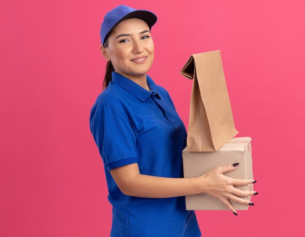 Glückliche junge Lieferfrau in der blauen Uniform und in der Kappe, die Papierpaket und Pappkarton hält, der vorne lächelnd zuversichtlich über rosa Wand steht