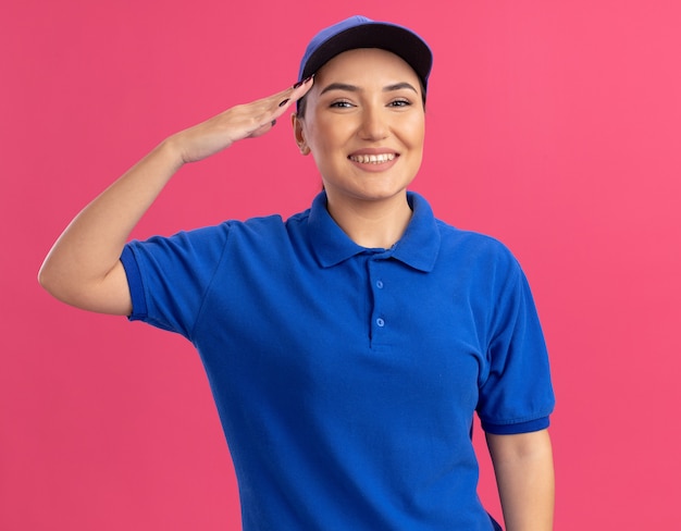 Glückliche junge Lieferfrau in der blauen Uniform und in der Kappe, die vorne lächelnd souverän salutierend über rosa Wand stehend schaut