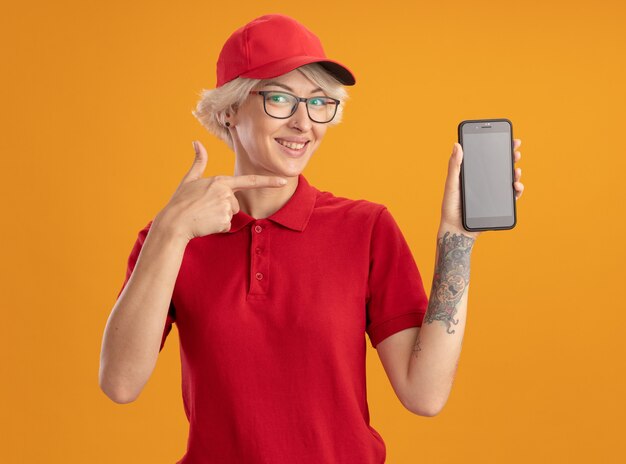 Glückliche junge Lieferfrau in der roten Uniform und in der Kappe, die die Brille trägt, die Smartphone zeigt, das mit Zeigefinger darauf zeigt und fröhlich über orange Wand steht