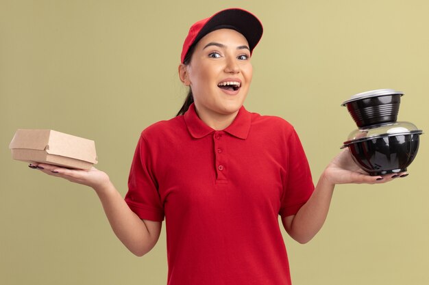 Glückliche junge Lieferfrau in der roten Uniform und in der Kappe, die Lebensmittelpakete halten, die vorne mit Lächeln auf Gesicht stehen über grüner Wand stehen