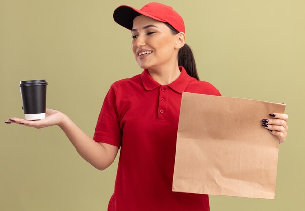 Glückliche junge Lieferfrau in der roten Uniform und in der Kappe, die Papierpaket hält, das Kaffeetasse mit Lächeln auf Gesicht über grüner Wand steht