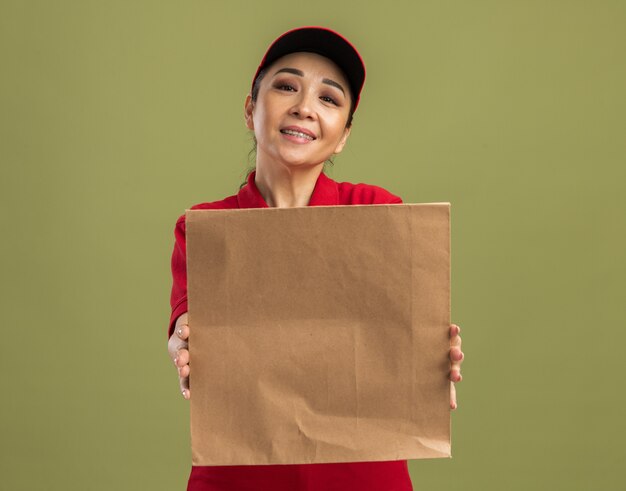 Glückliche junge Lieferfrau in roter Uniform und Mütze, die Papierpaket mit einem Lächeln im Gesicht hält