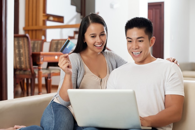 Glückliche junge Mischrassepaare mit dem Laptop, der auf Sofa sitzt und online kauft