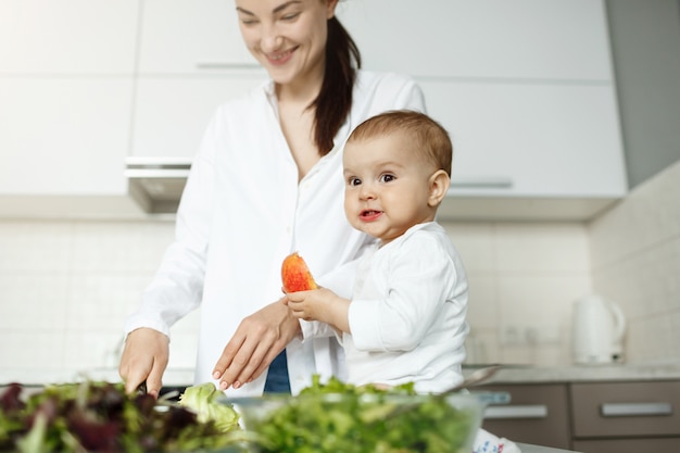 Glückliche junge Mutter, die Frühstück in heller Küche mit ihrem kleinen süßen Sohn kocht. Kind, das Pfirsich mit lustigem Ausdruck isst, während Mutter arbeitet.