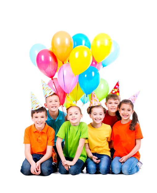 Glückliche Jungen und Mädchen im Partyhut mit farbigen Luftballons, die auf dem Boden sitzen - lokalisiert auf Weiß