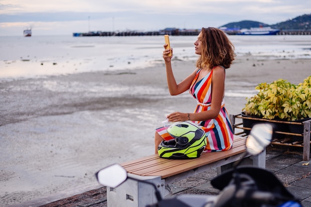 Glückliche kaukasische Bikerfrau im bunten Sommerkleid im Urlaub mit Motorradhelm
