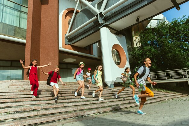 Glückliche Kinder, die an der Straße der Stadt am sonnigen Sommertag vor dem modernen Gebäude spielen. Gruppe von glücklichen Kindern oder Jugendlichen, die Spaß zusammen haben. Konzept der Freundschaft, Kindheit, Sommer, Ferien.