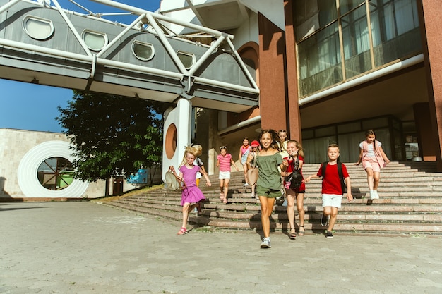 Glückliche Kinder, die an der Straße der Stadt am sonnigen Sommertag vor dem modernen Gebäude spielen. Gruppe von glücklichen Kindern oder Jugendlichen, die Spaß zusammen haben. Konzept der Freundschaft, Kindheit, Sommer, Ferien.