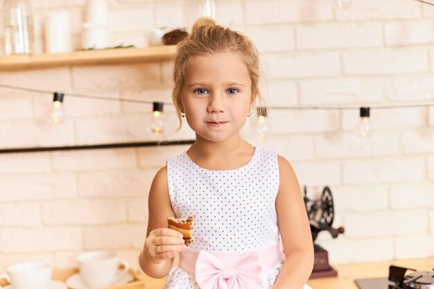 Glückliche Kindheit, Spaß und Freude Konzept. Innenaufnahme des süßen entzückenden Babymädchens, das schönes Kleid trägt, das am Esstisch im stilvollen Kücheninnenraum sitzt, lacht, köstlichen Keks oder Kuchen kaut