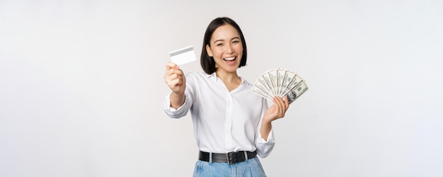 Glückliche Koreanerin, die Kreditkarte und Gelddollar hält, lächelt und lacht und posiert vor weißem Studiohintergrund