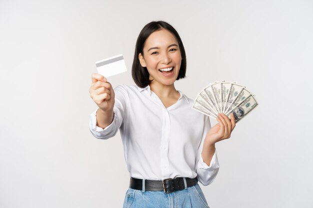 Glückliche Koreanerin, die Kreditkarte und Gelddollar hält, lächelt und lacht und posiert vor weißem Studiohintergrund