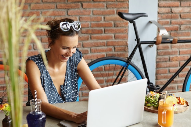 Glückliche lächelnde Frau, die Freunde online in den sozialen Medien mitteilt, im Internet surft, kostenloses WLAN auf ihrem modernen Laptop verwendet und mit Essen am Tisch sitzt. Menschen