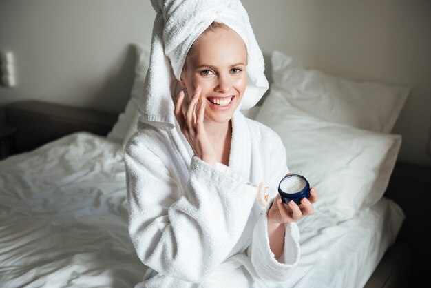 Glückliche lächelnde Frau im Bademantel, die Creme auf Gesicht setzt