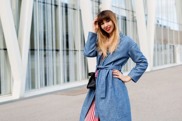 Glückliche lächelnde Frau im modischen blauen Mantel und im gestreiften roten Kleid, die auf modernem Geschäftszentrum gehen