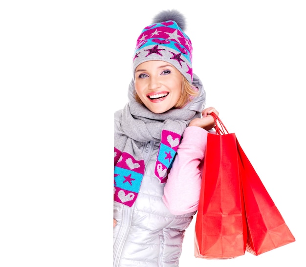 Glückliche lächelnde Frau mit Geschenken nach dem Einkaufen schaut aus dem weißen leeren Plakat heraus