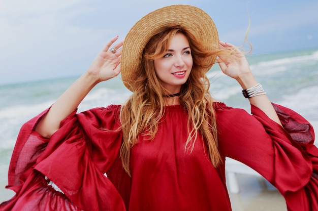 Glückliche lächelnde weiße Ingwerfrau im stilvollen roten Kleid, das Selbstporträt-Handy am Strand nahe Ozean macht. Strohhut tragen.