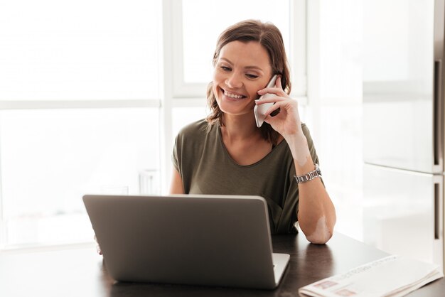 Glückliche lässige Frau, die durch Smartphone spricht, während sie nahe dem Tisch mit Laptop-Computer sitzt