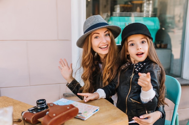 Glückliche liebende Familie. Mutter und ihre Tochter sitzen in einem Stadtcafé und schauen überrascht in die Kamera und das Mädchen, das den Weg zeigt. Auf dem Tisch liegen eine Karte und Kameras. Wahre Gefühle, gute Laune ..