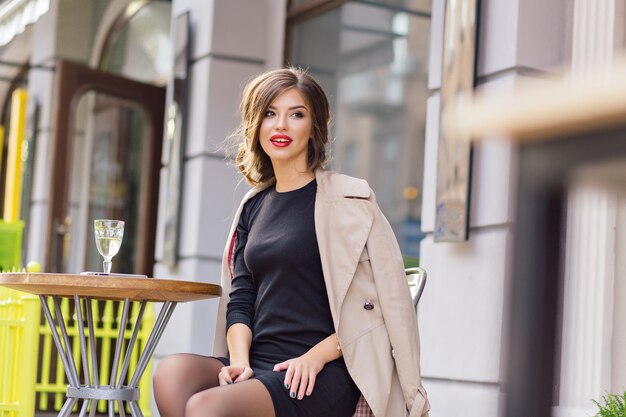 Glückliche liebenswerte Frau im schwarzen Kleid und im beigen Mantel, die in der Cafeteria im Freien sitzen und mit einem Glas Wein ruhen