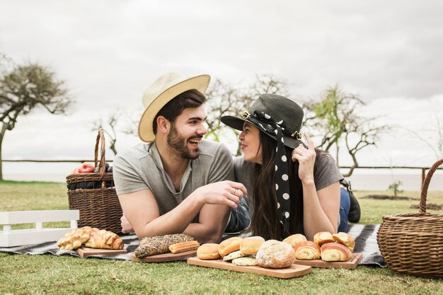 Glückliche liebevolle junge Paare, die auf Decke mit gebackenen Broten am Picknick liegen