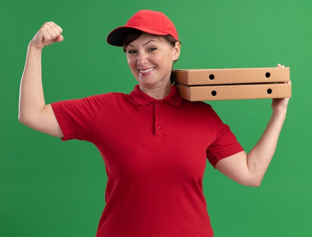 Glückliche Lieferfrau mittleren Alters in der roten Uniform und in der Kappe, die Pizzakästen hält, die Faust betrachten, die vorne lächelnd zuversichtlich über grüner Wand steht