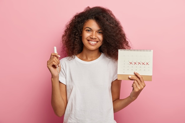 Glückliche lockige haarige Dame hält Menstruationskalender mit markierten PMS-Tagen und Tampon, gekleidet in lässigem weißem T-Shirt, lokalisiert über rosa Hintergrund